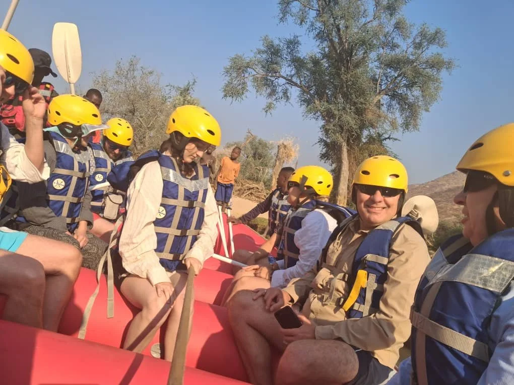 People wearing yellow helmets and life jackets riding in a red inflatable boat in a dry, desert landscape with trees.