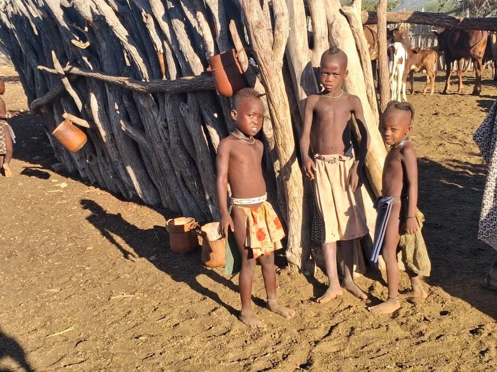 Three children standing near a wooden structure made of logs, with horses and other children in the background.