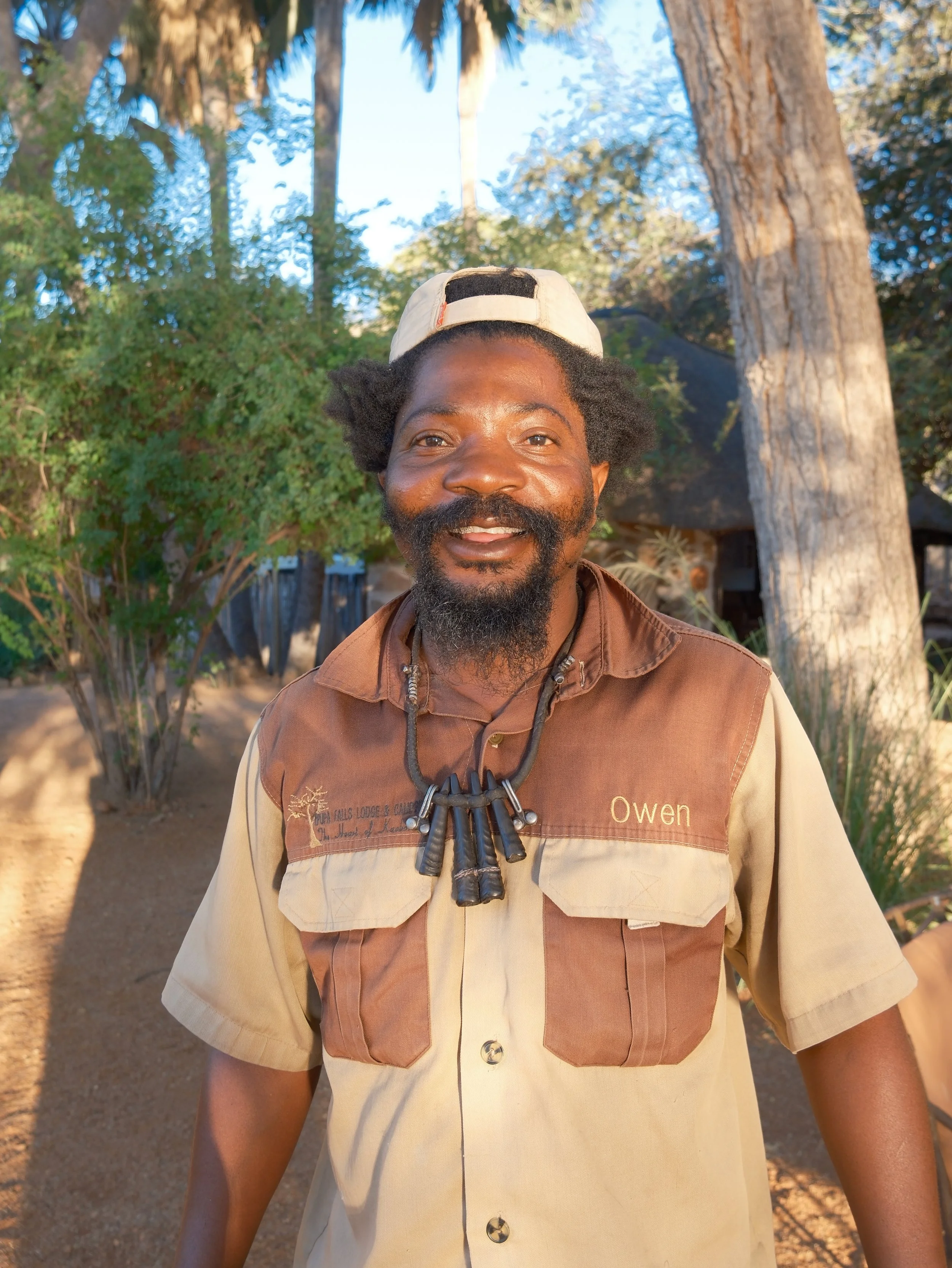 A smiling man with a beard, wearing a tan and brown shirt with a name tag that says Owen, a necklace with several black tubes, and a backward white baseball cap, standing outside with trees and a blue sky in the background.