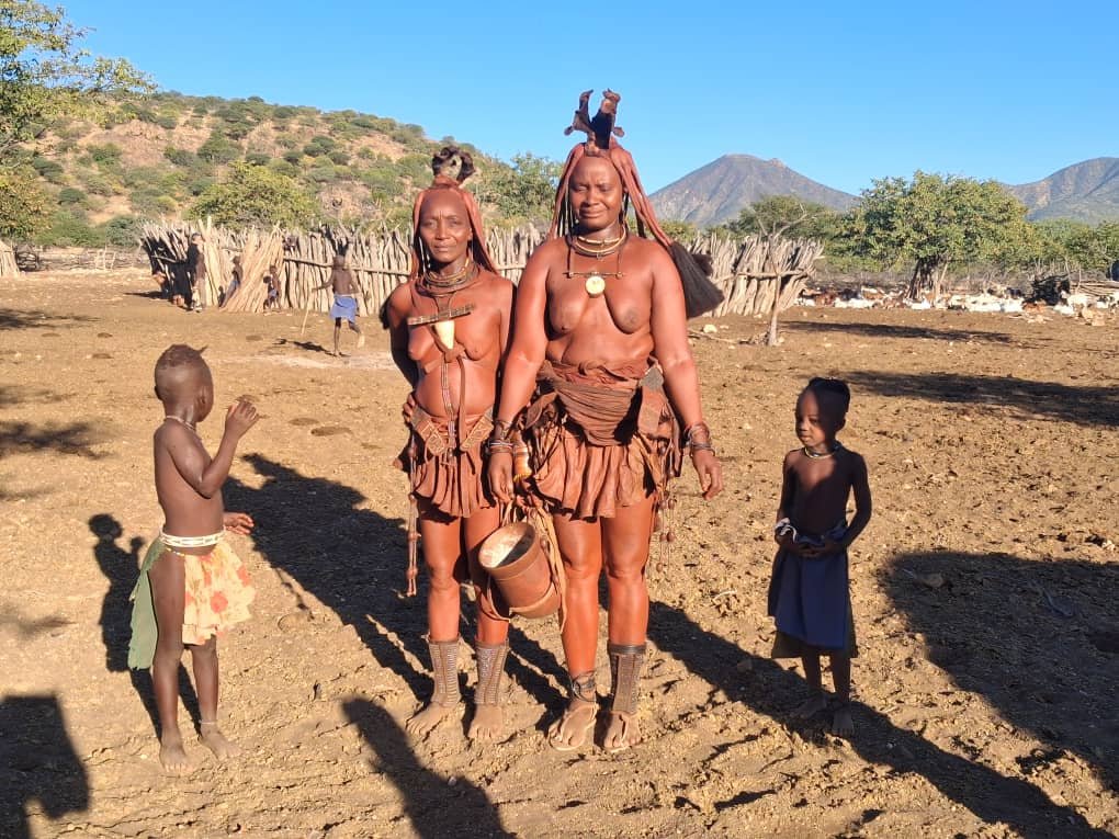 Two indigenous women with traditional attire and jewelry, standing outdoors on dirt ground, with two children nearby. The background shows a fence, sparse trees, and mountains under a clear blue sky.