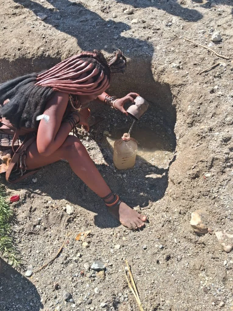 A person with long, pink dreadlocks wearing traditional jewelry and clothing kneels in a dug hole in the ground, pouring water from a small cup into a plastic bottle.