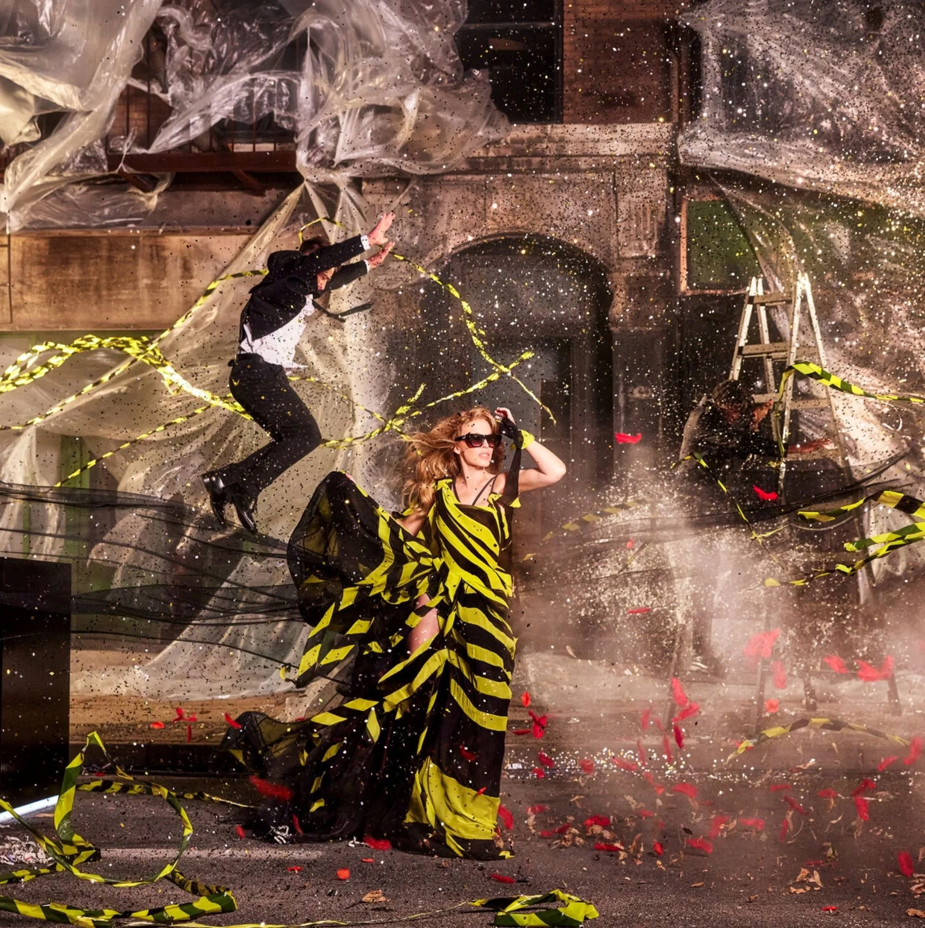 Kylie Minogue People celebrating with confetti and streamers, with yellow and black striped ribbons, in front of a brick building with archways, in a festive atmosphere.