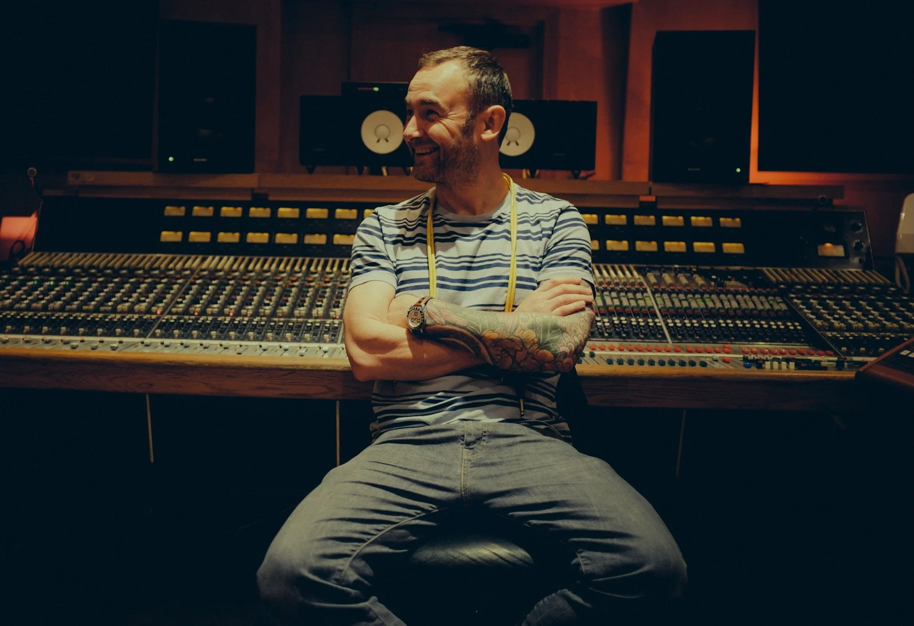 A man with tattoos on his left arm, wearing a striped t-shirt and gray pants, smiling and sitting in front of a large audio mixing console in a recording studio.