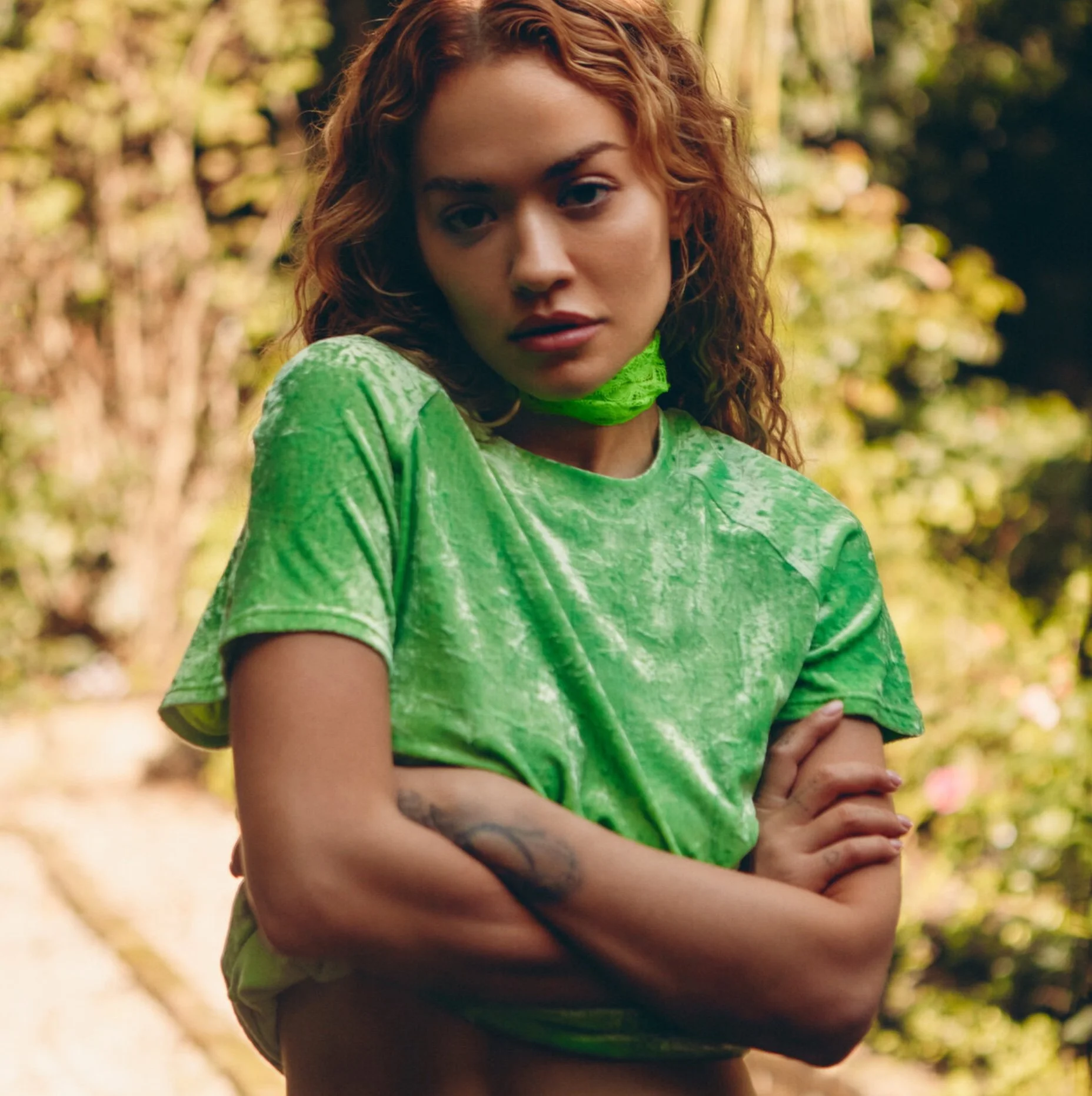 A young woman with curly red hair, wearing a green tie-dye shirt, standing outdoors in a wooded area with sunlight filtering through the trees.