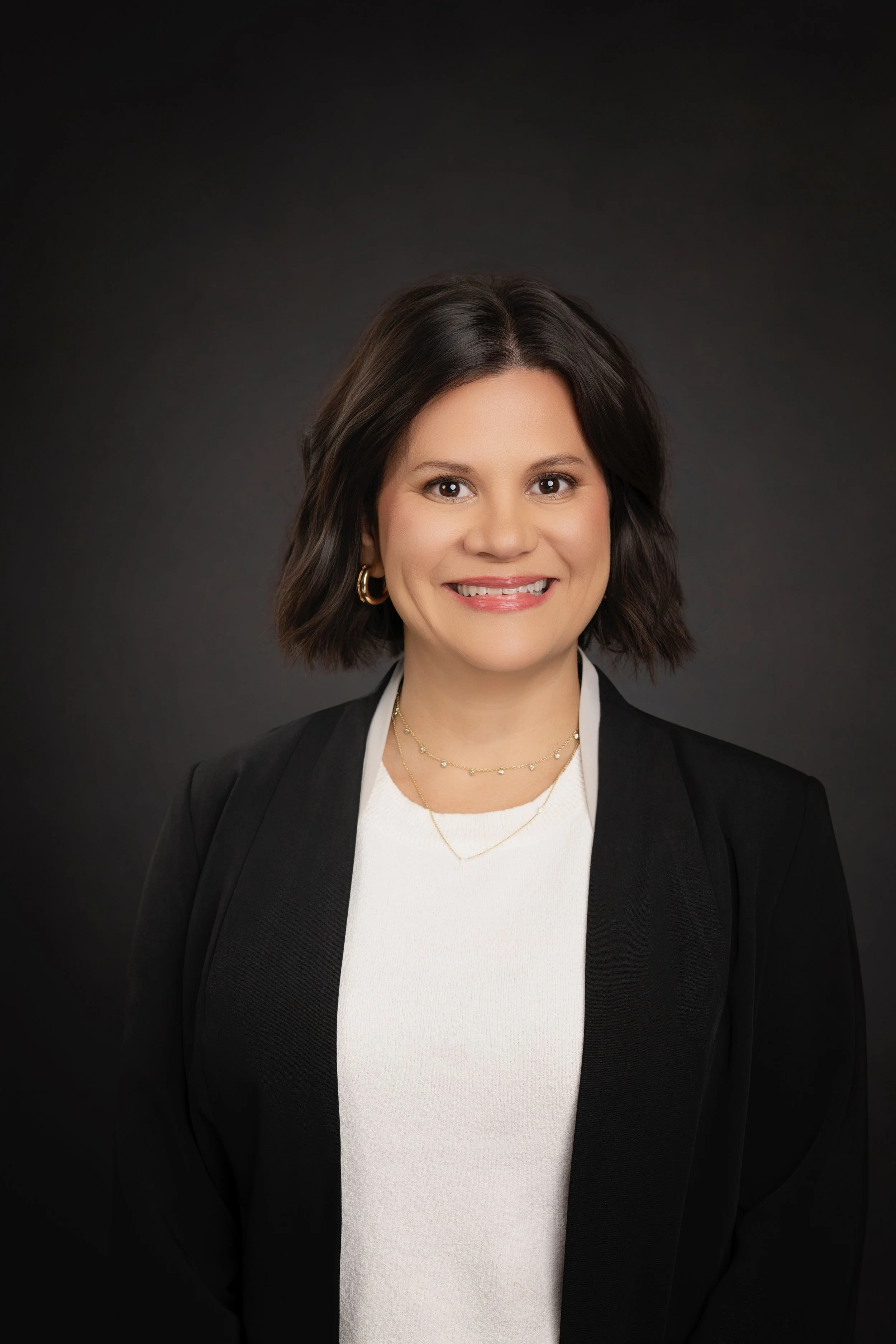 A woman with short dark hair, wearing a black blazer, white top, gold hoop earrings, and layered gold necklaces, smiling against a dark background.