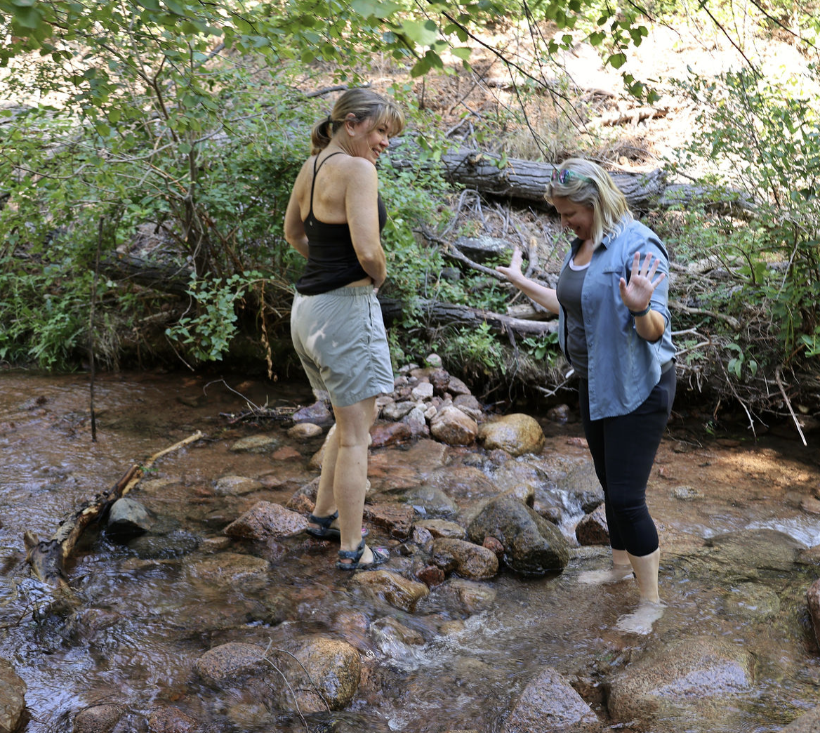Two women over 50 sharing an adventure in a shallow creek surrounded by green foliage and rocks, smiling and engaging in a playful conversation.