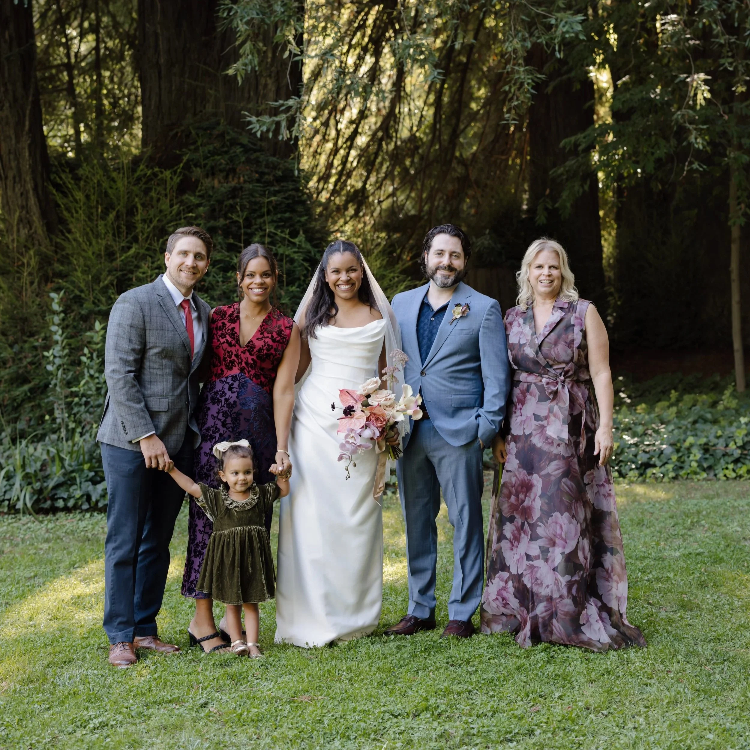 Group of six people standing outside in front of trees, including a newlywed bride and groom, two men, and two women, with two young girls in the front.