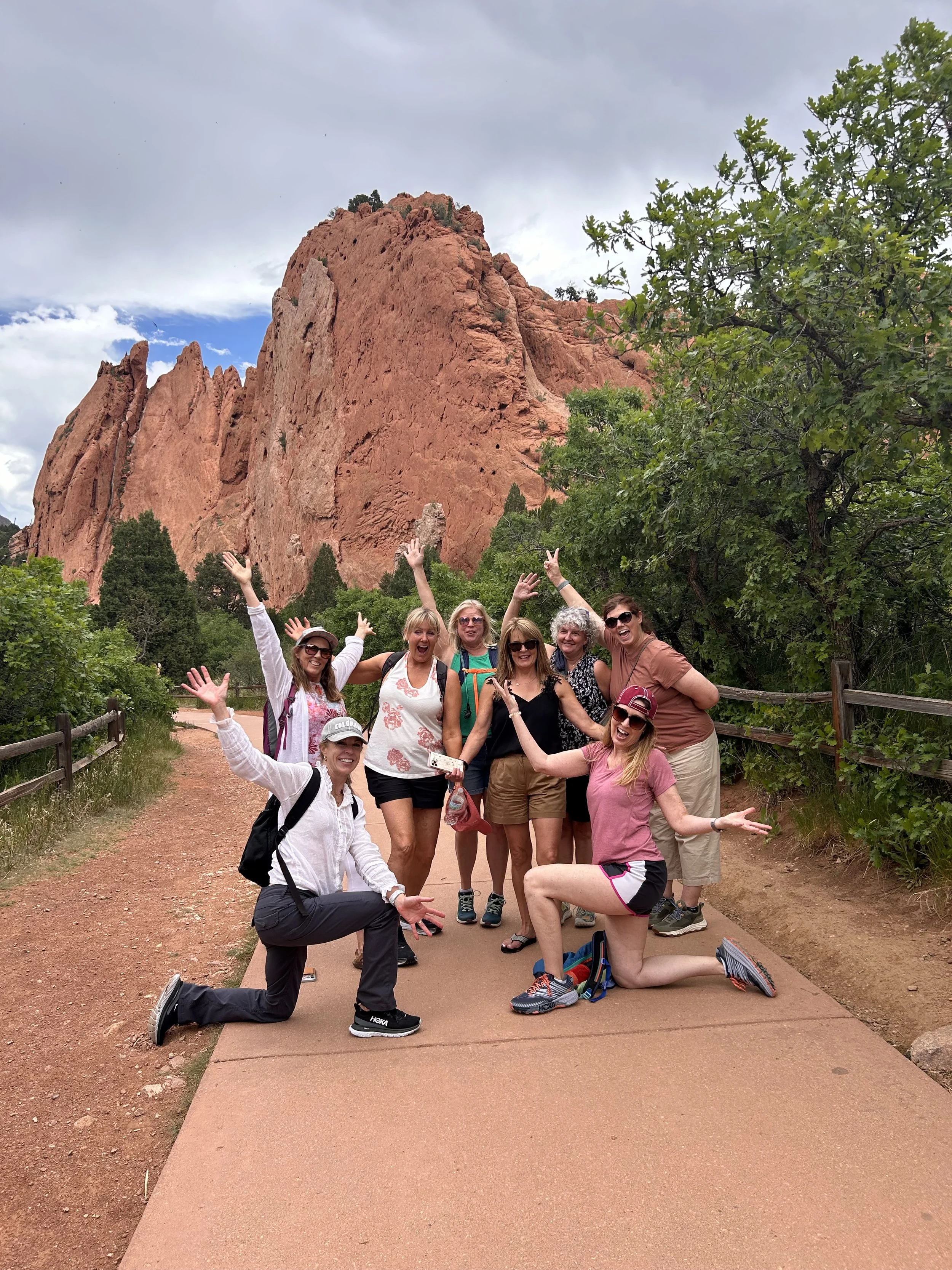 Group of 50+women  outdoors at a park near red rock formations, some standing and some kneeling, smiling and making hand gestures. Enjoying each others company and feeling alive