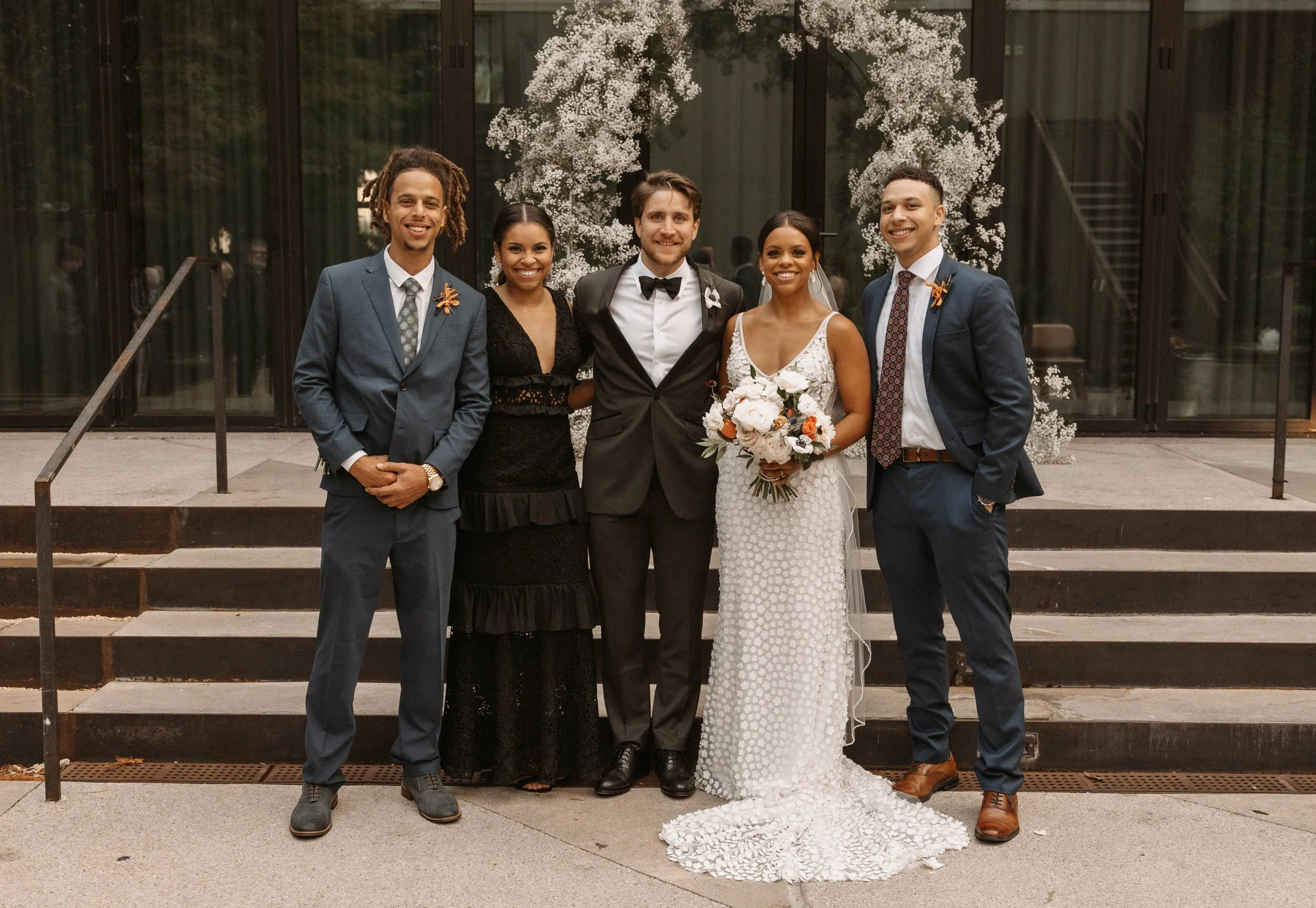 Group of five people at a wedding, with a bride in a white wedding dress holding a bouquet of flowers, standing in front of a building with glass doors and white floral decorations.