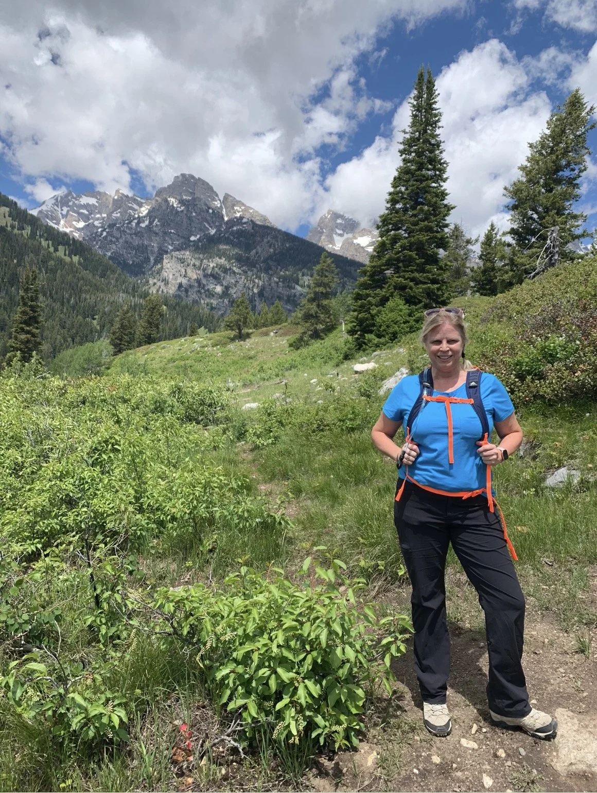 A woman hiking in a lush green mountain landscape with tall trees and snowy mountain peaks in the background, under a partly cloudy sky.