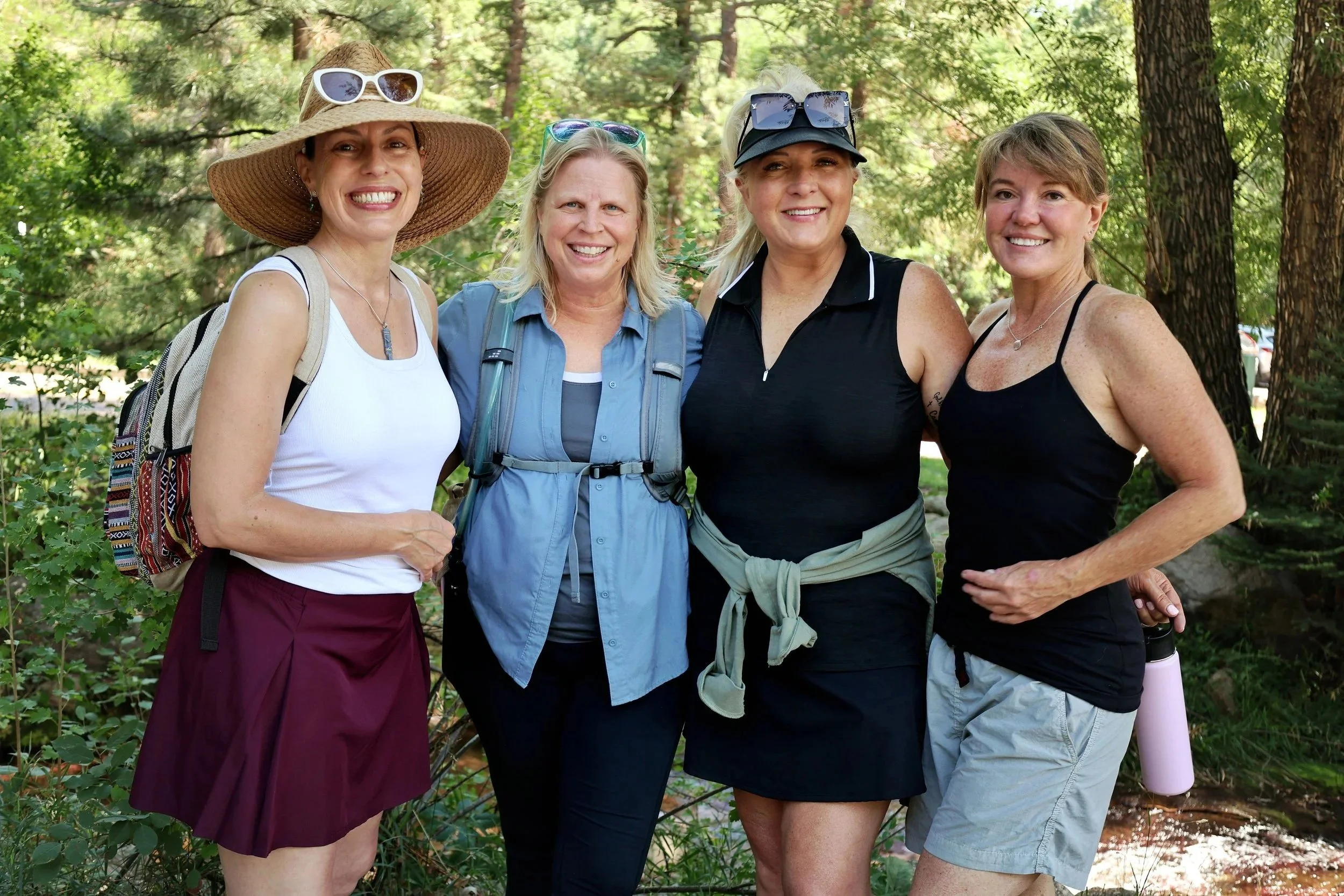 Four women ovef 50 standing together outdoors in a wooded area, smiling at the camera, dressed in casual and outdoor hiking attire. enjoying nature walk and good conversation