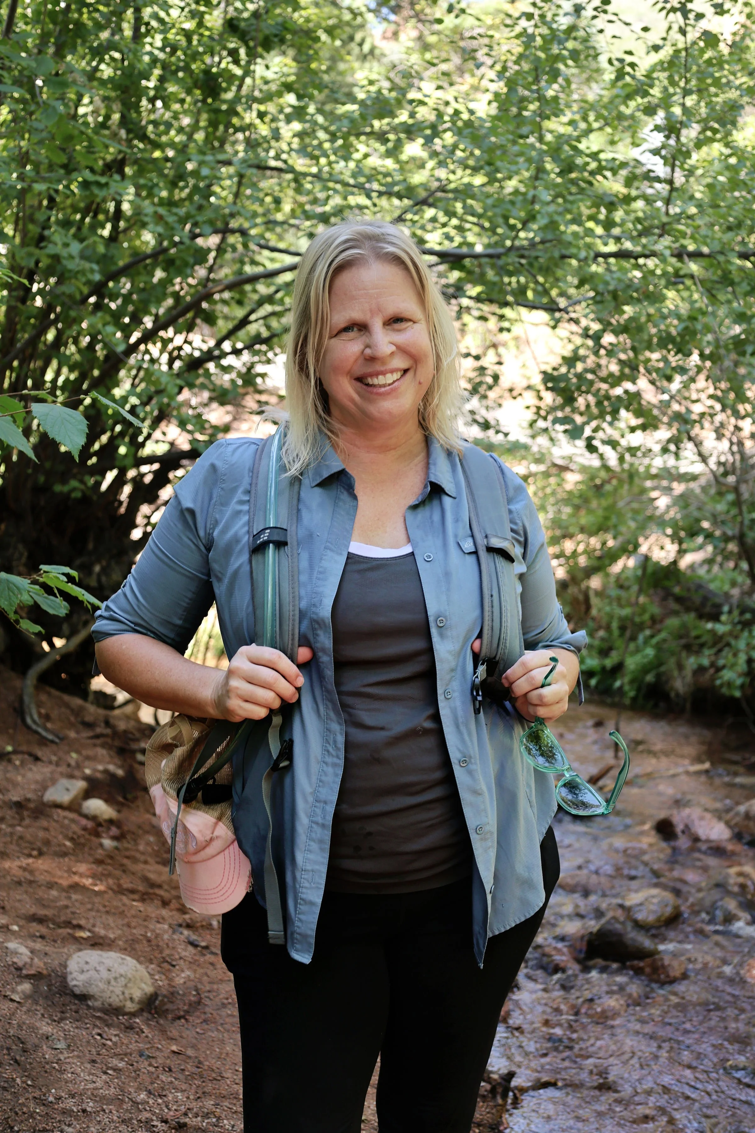 A woman with blonde hair smiling and standing outdoors in a wooded area, holding a pair of sunglasses and wearing a blue jacket over a dark shirt, with a backpack and a pink hat hanging from her backpack strap.