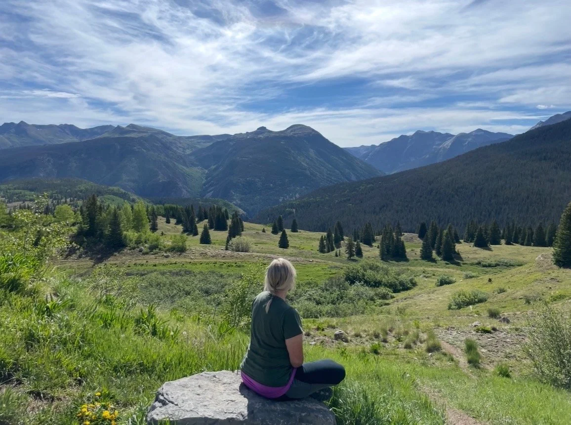 A woman sitting on a rock in a lush green mountain valley with pine trees and mountains in the background, under a partly cloudy sky. feeling the peacefullness of the outdoors and nature healing