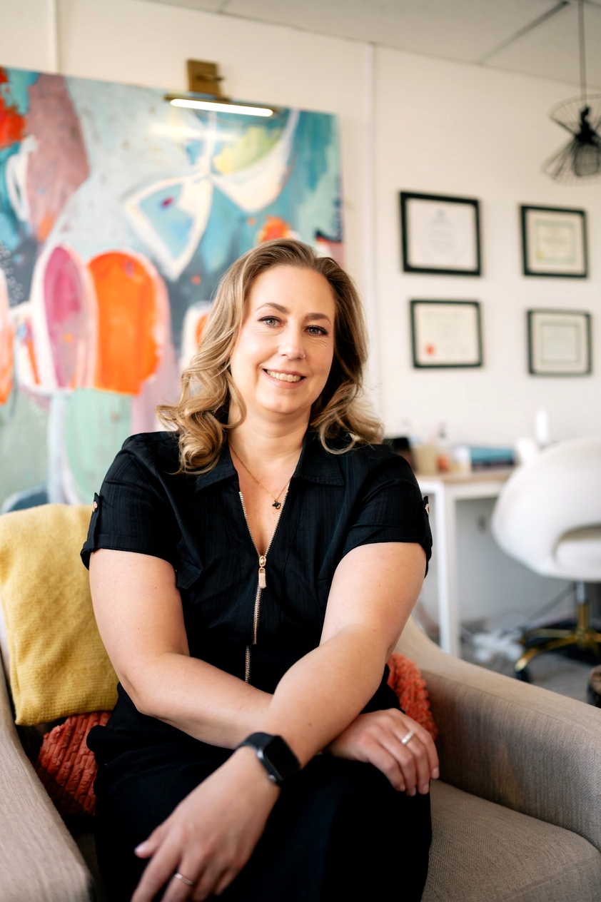 A woman with curly blonde hair smiling and sitting on a beige couch in an art studio or office, with colorful abstract artwork on the wall behind her and framed certificates on the wall to the right.