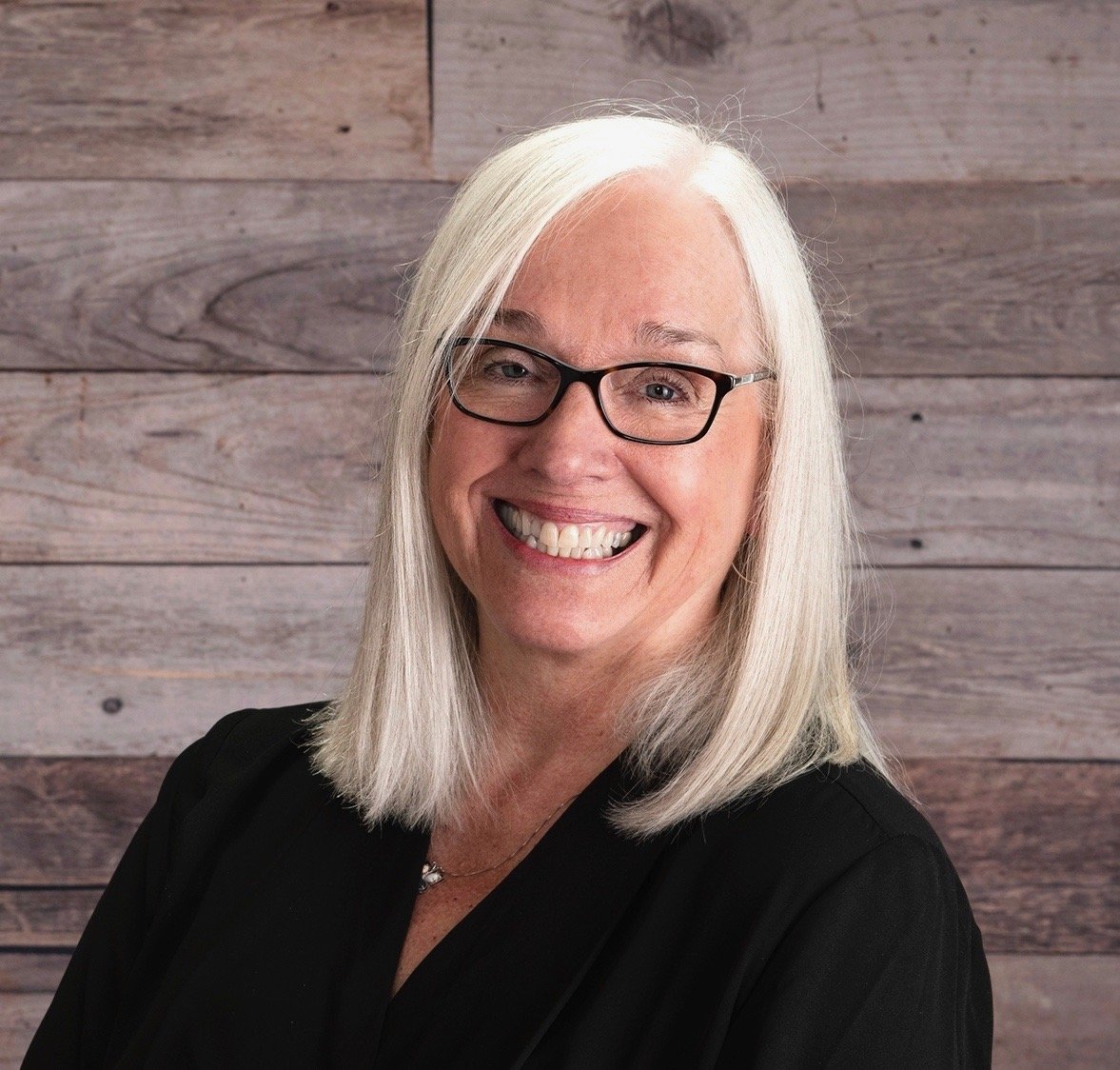 A smiling middle-aged woman with long white hair and glasses, wearing a black top, standing in front of a wooden wall.