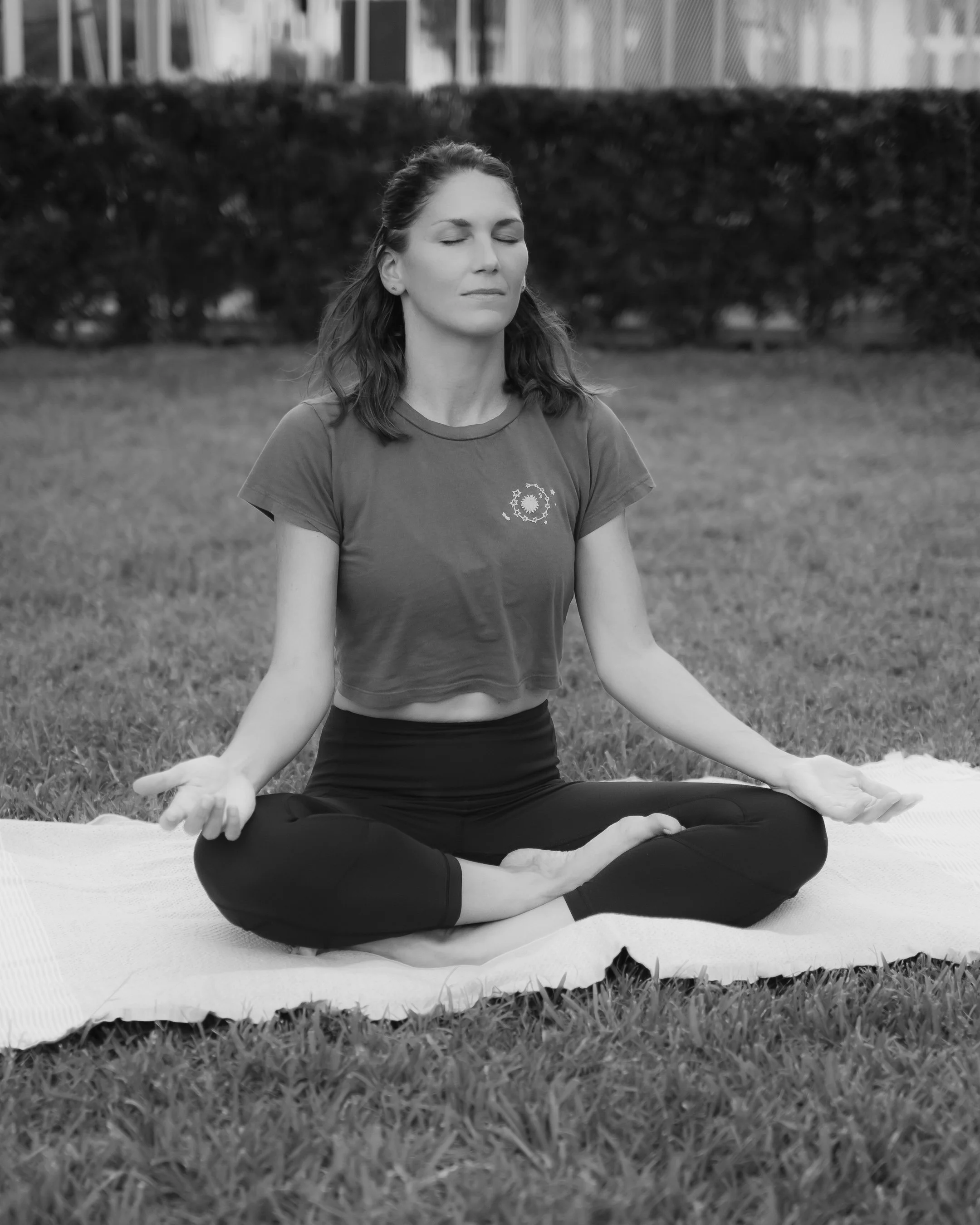 A woman practicing yoga outdoors on a grassy field, sitting cross-legged with her hands resting on her knees and eyes closed in meditation.