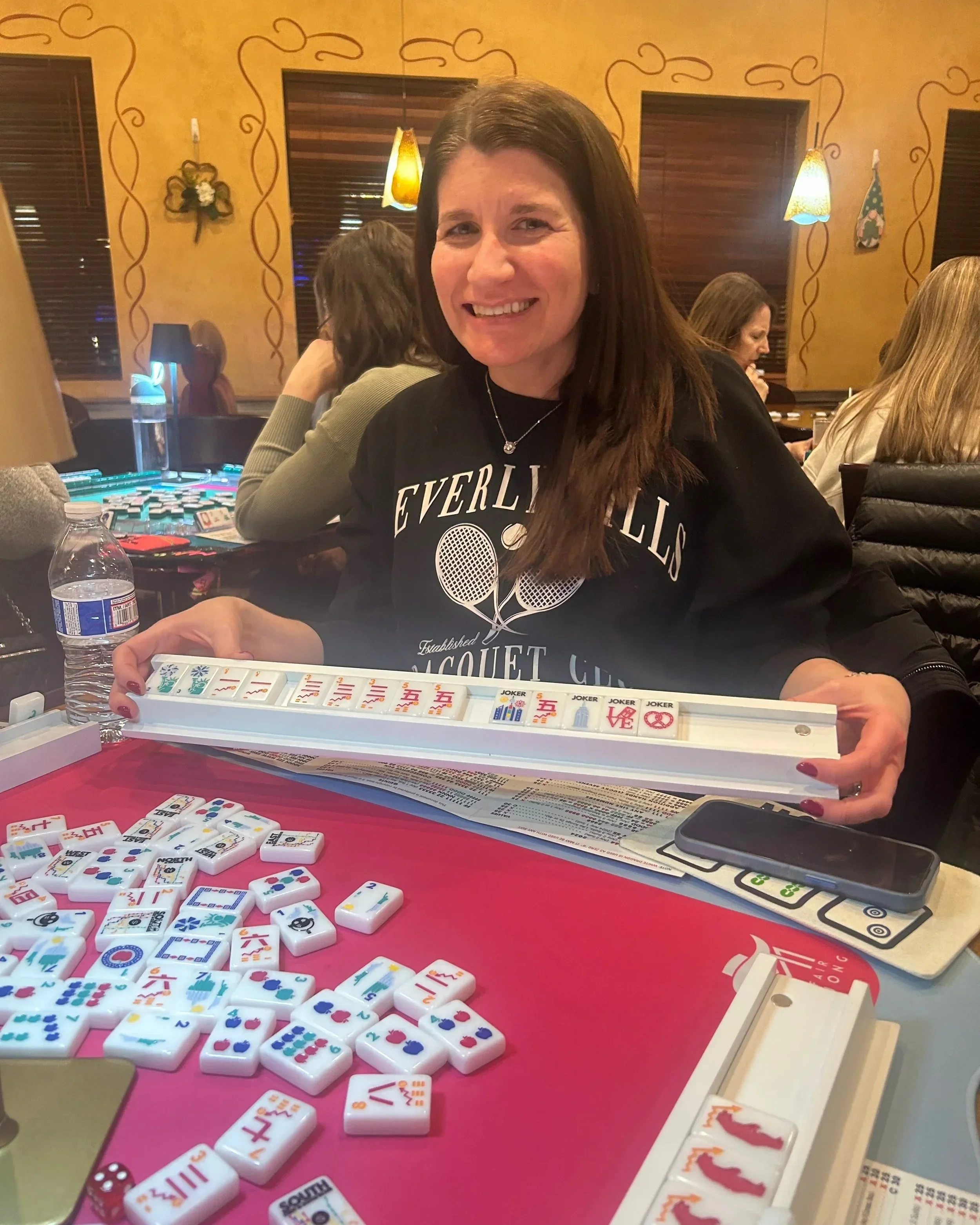 A woman with long dark hair smiling while playing Mahjong at a table in a restaurant. The table has Mahjong tiles and a bottle of water. Other people are sitting at nearby tables.
