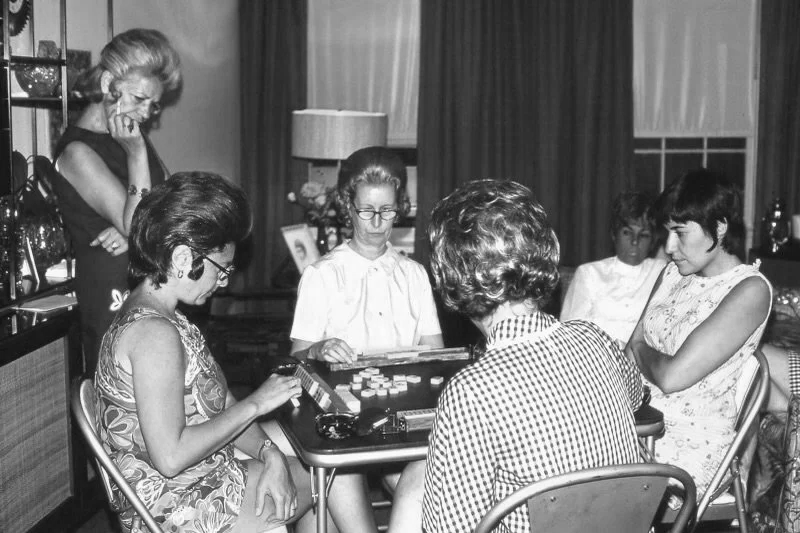 Six women gathered around a table playing a game, with one standing and five seated, in a decorated room with curtains and lamps.
