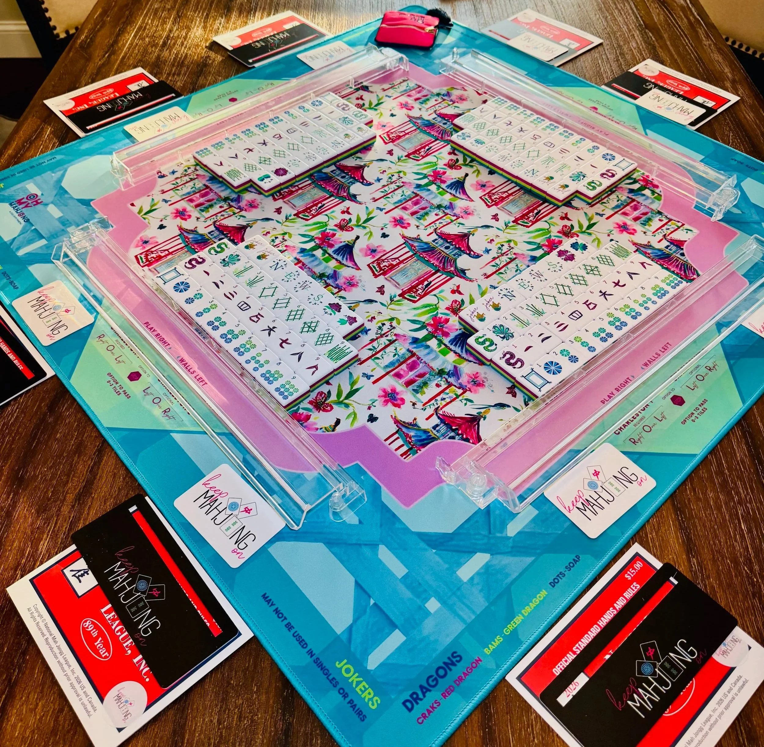 A Mahjong game set on a wooden table includes a blue plastic mat, colorful tiles, and various instructions and rule sheets.