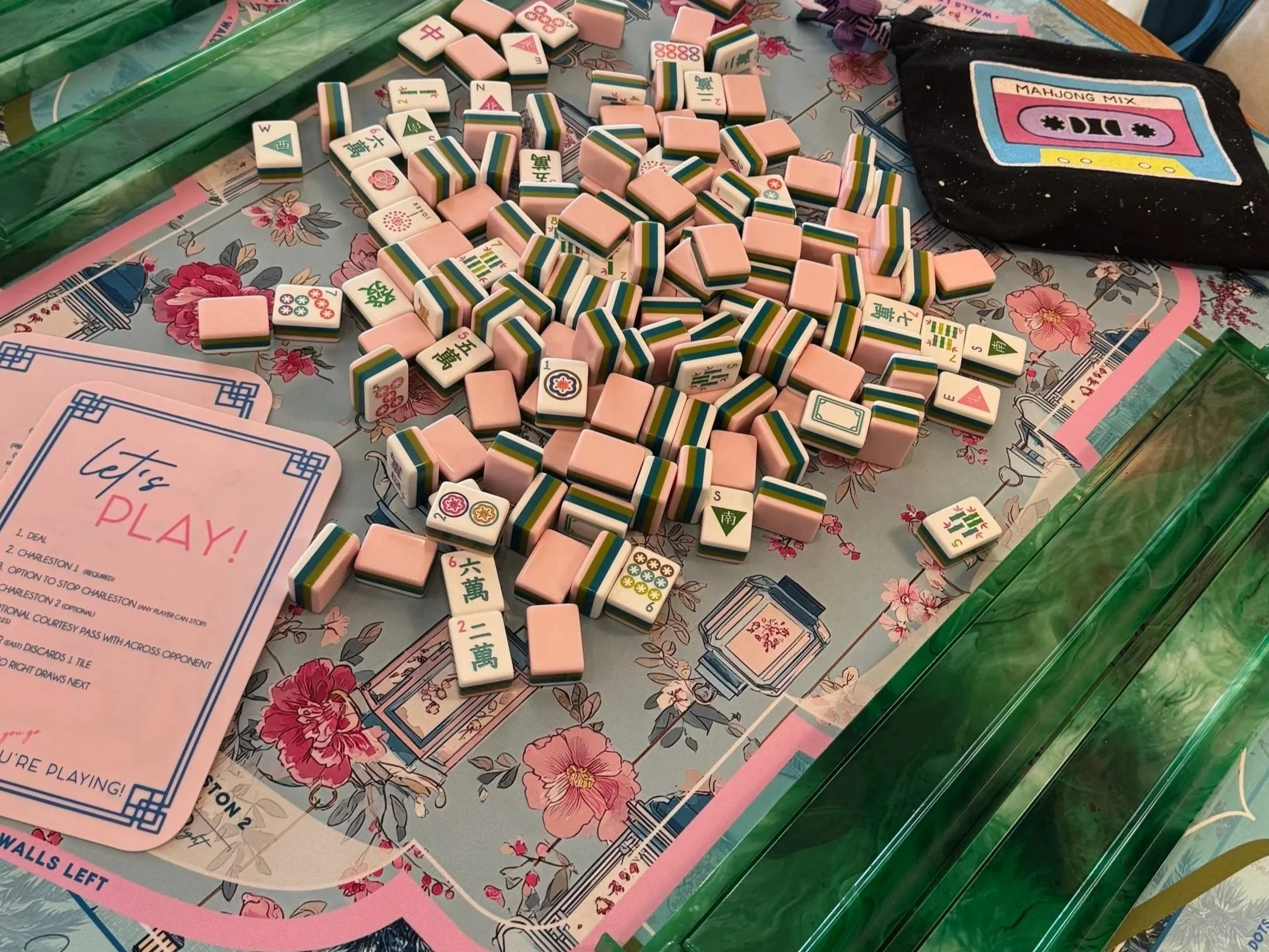 A mahjong game in progress with tiles scattered on a floral tablecloth, with a black bag labeled 'MAHJONG MIX' nearby, and some pink and green domino tiles.
