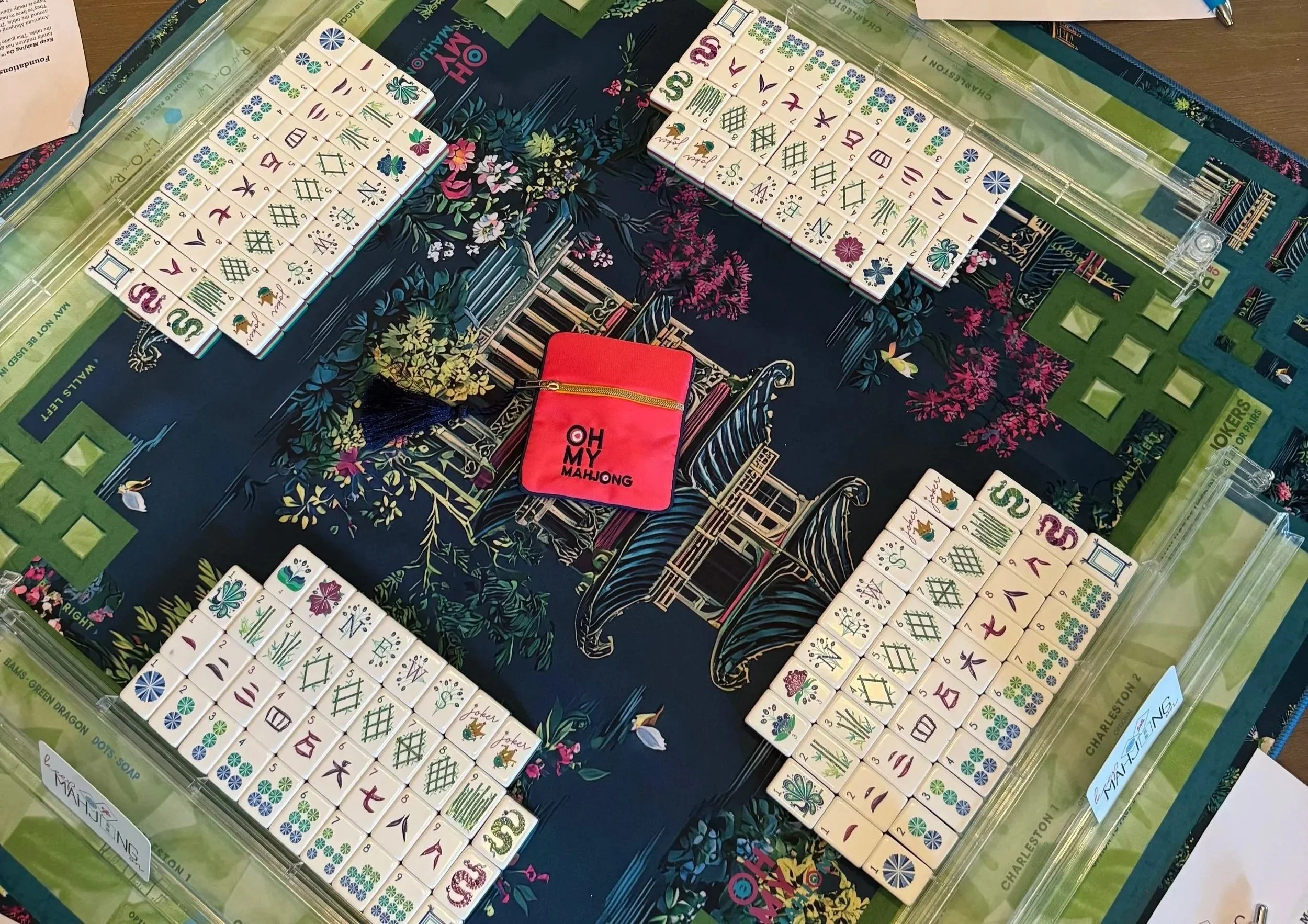 A mahjong game in progress with sets of tiles arranged on a floral-patterned table. The tiles feature various symbols, numbers, and designs, with some tiles forming groups. There is a red pouch labeled 'OH MY MAHJONG' near the center of the table.