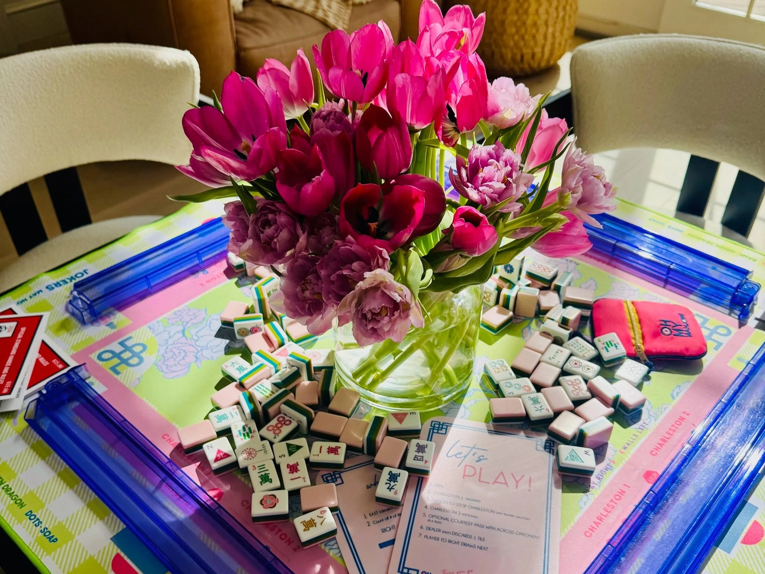 A colorful bouquet of pink tulips and other flowers in a glass vase on top of a mahjong game with scattered mahjong tiles around. The background includes two chairs and a basket in a cozy room setting.
