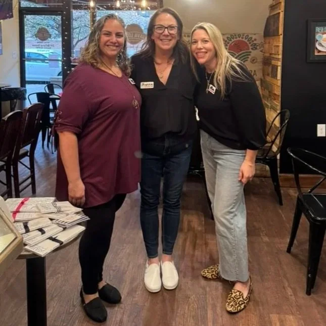 Three women standing together inside a restaurant, smiling at the camera. They are dressed casually, and there are tables and chairs around them. One woman is wearing a maroon top, another in black, and the third in light-colored pants with leopard-print shoes.