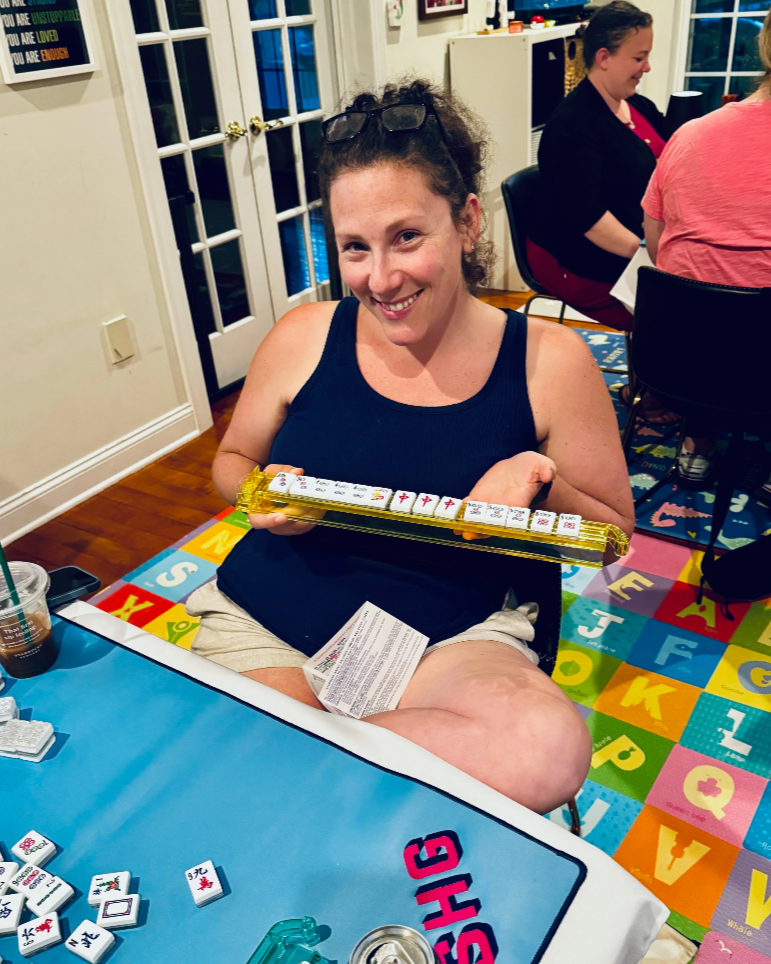 Woman smiling at the camera while holding a Mahjong tile rack with Mahjong tiles. She is sitting at a table with Mahjong tiles and a drink cup. Other people are in the background in a brightly decorated room with colorful alphabet carpet.