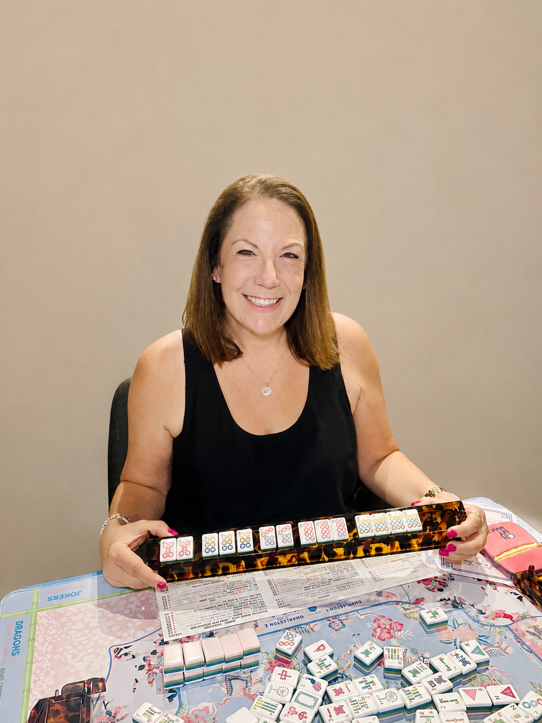 A woman smiling at the camera while playing mahjong, holding mahjong tiles in her hands, sitting at a table with mahjong tiles and a game board.