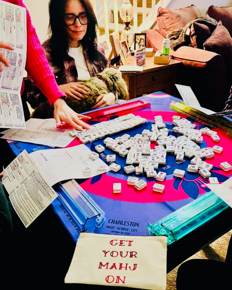 Group of people playing a game of Mahjong at a table covered with game tiles, manuals, and score sheets in a living room.