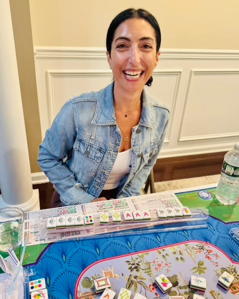 Woman sitting at a table playing Mahjong, smiling, with tiles in front of her and a water bottle on the side.