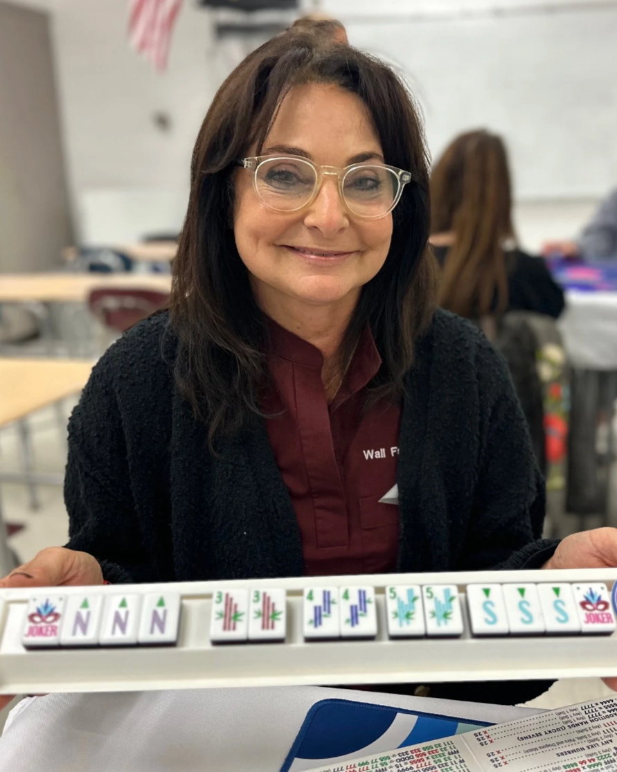 A woman with shoulder-length dark hair, glasses, and a maroon shirt with a black cardigan is sitting at a table in a classroom, smiling at the camera. Behind her, other people are seated at tables.