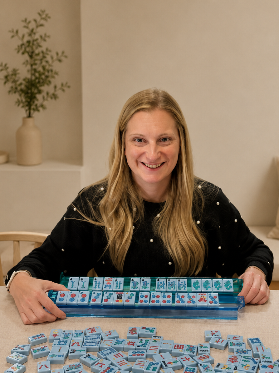 Woman smiling at a table with Mahjong tiles, some arranged in a rack and others scattered on the table.