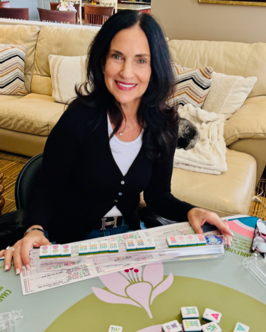 Woman with dark hair smiling at a table with Mahjong tiles, seated in a living room with a beige couch and pillows in the background.