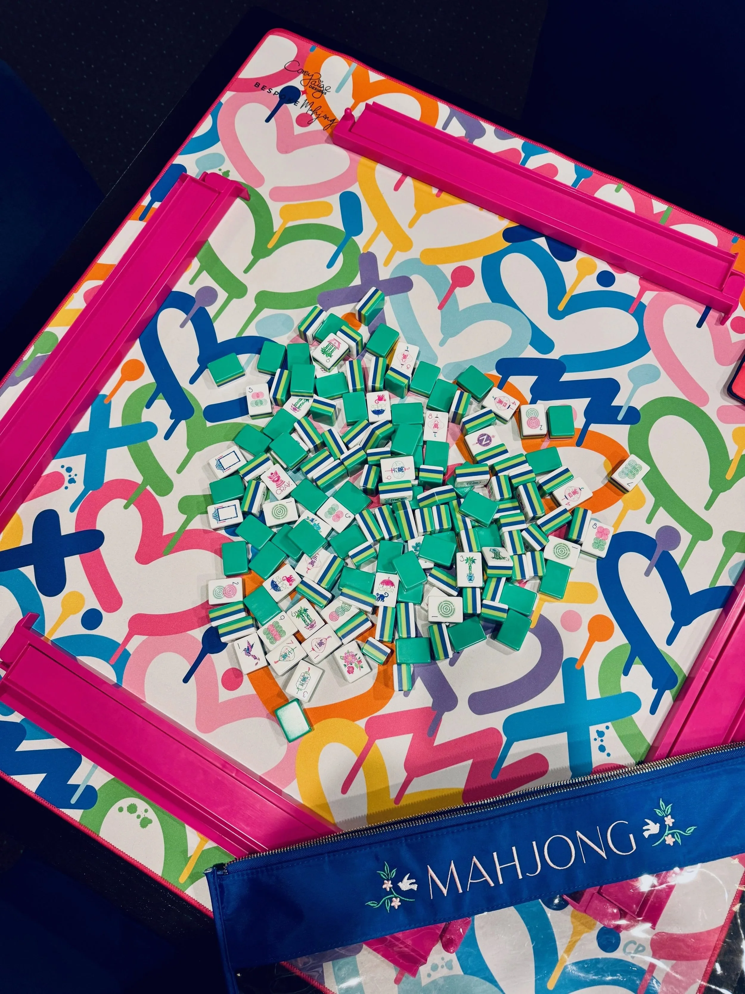 A colorful Mahjong game set with green, white, and striped tiles on a vibrant, multicolored table with a pink and blue border.