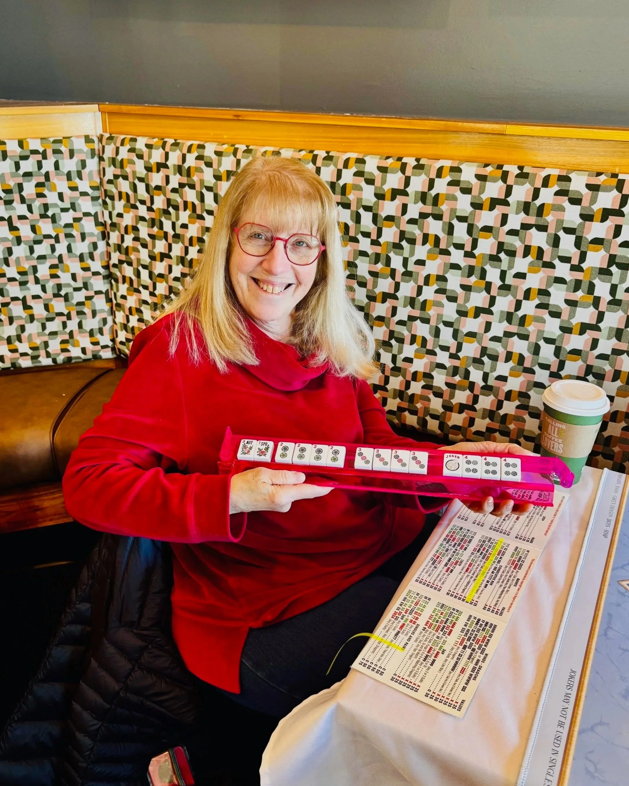 A smiling woman with red glasses and blonde hair sitting at a restaurant table, holding a bingo card, with a drink on the table, and a menu in front of her.