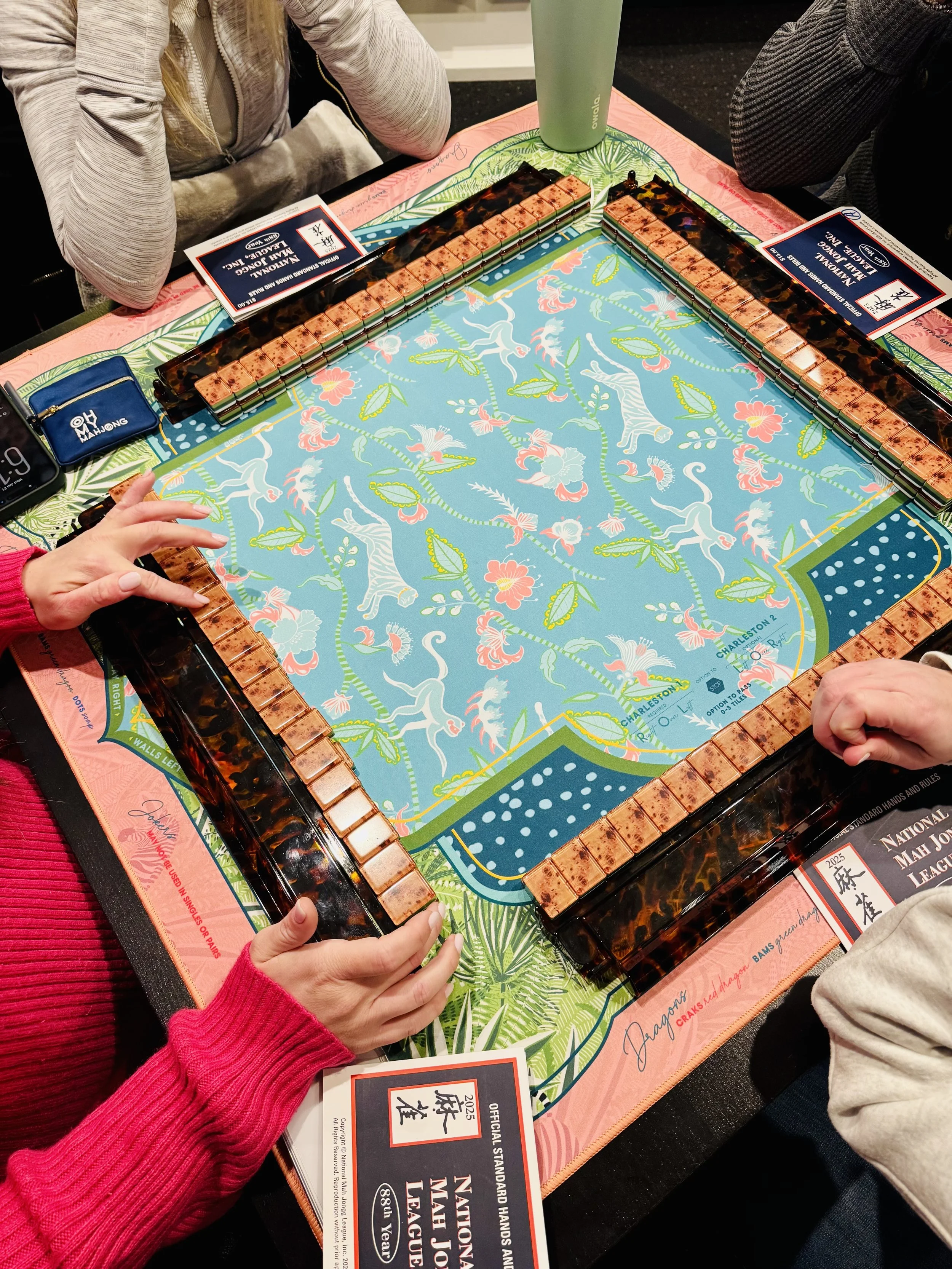 A mahjong game being played on a colorful table with two players holding hands, and a set of mahjong tiles arranged on the table. There are additional items such as a green tumbler, a blue wallet, and game rules visible on the table.