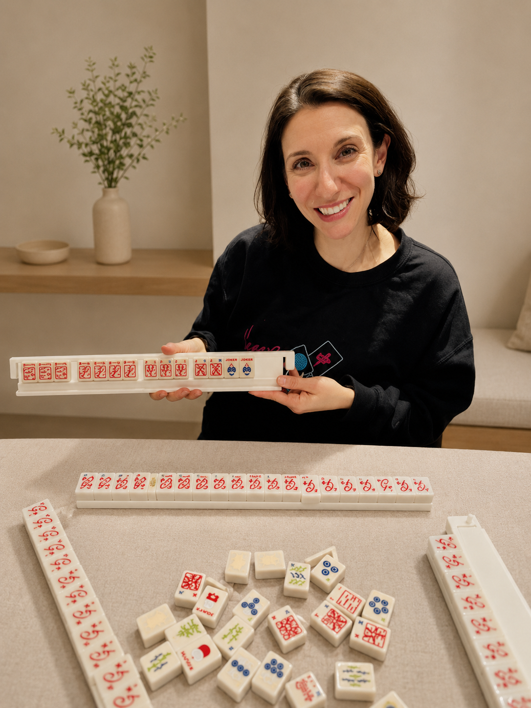 Woman playing mahjong with tiles arranged on the table and a rack in her hands, smiling at the camera.