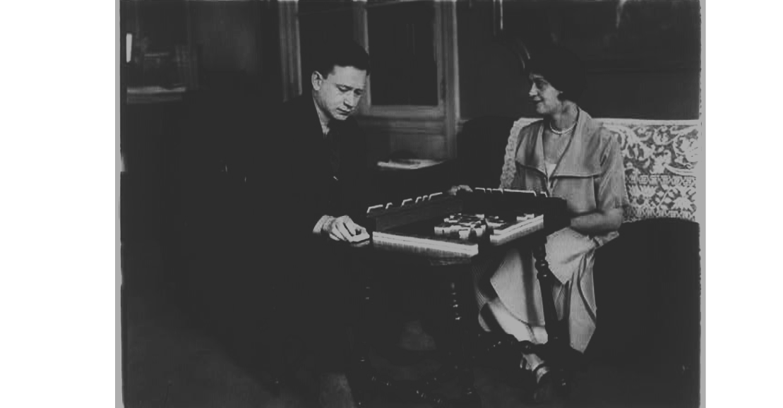 A black-and-white photo of a man and woman playing mahjong at a table in a cozy room with wood-paneled walls.