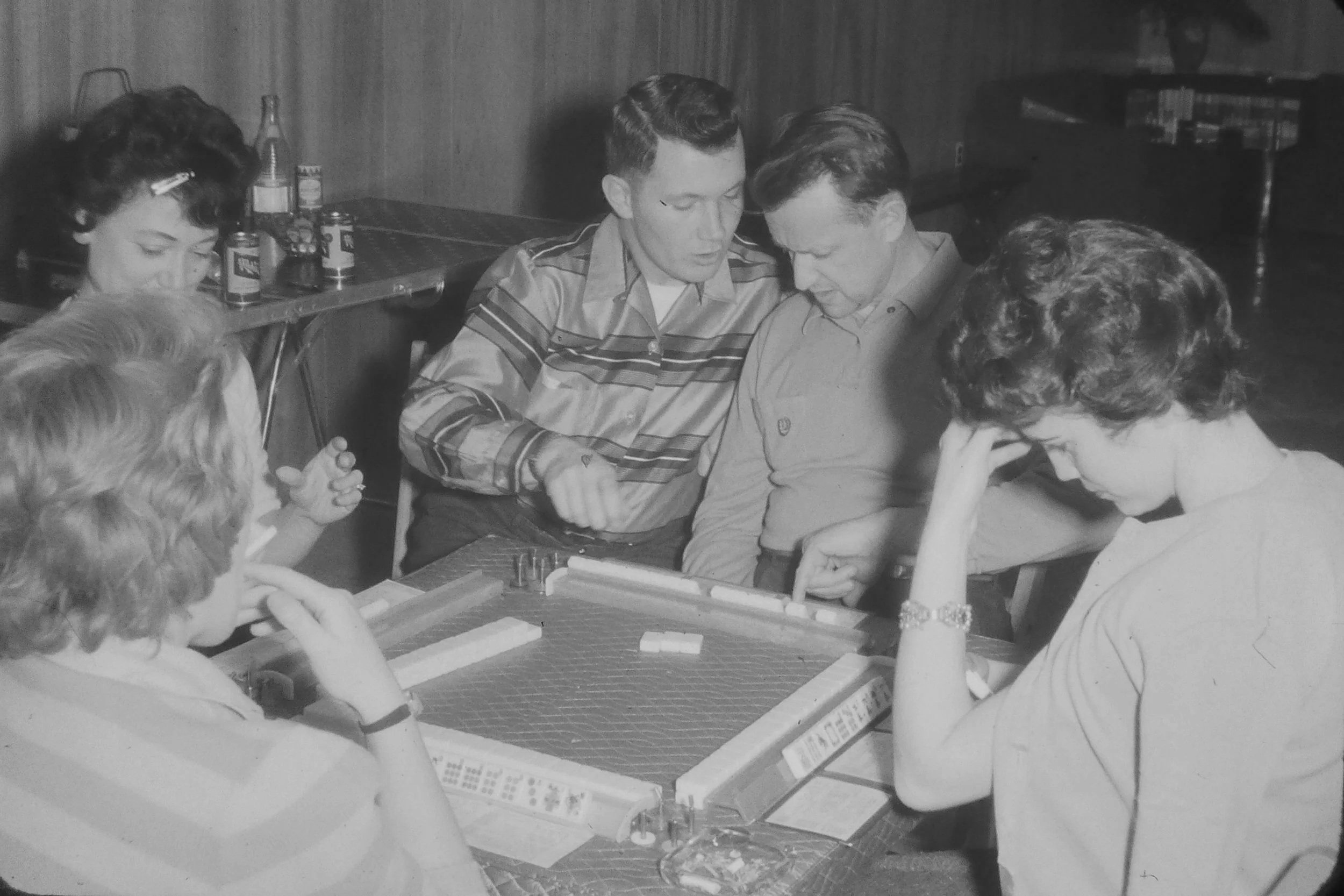 Black and white photo of five women and two men gathered around a table playing mahjong.