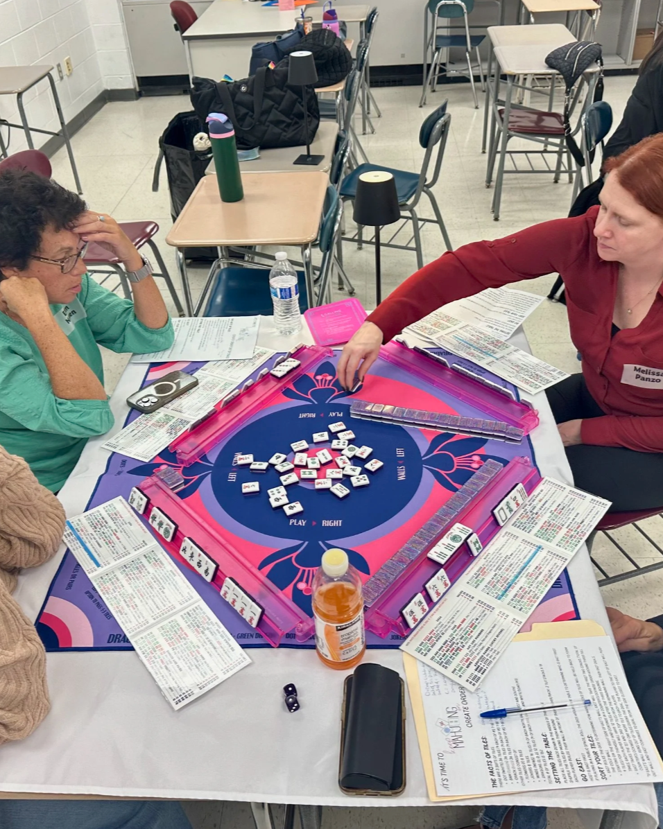 People playing a game of Mahjong at a table surrounded by papers, drinks, and other items in a classroom or meeting room setting.