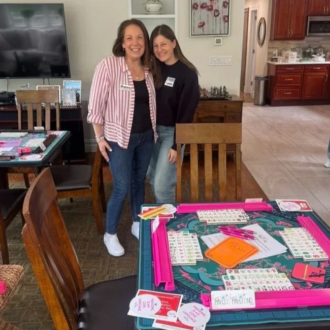 Two women smiling and standing inside a home, with a table set up for a game or activity, and a kitchen visible in the background.
