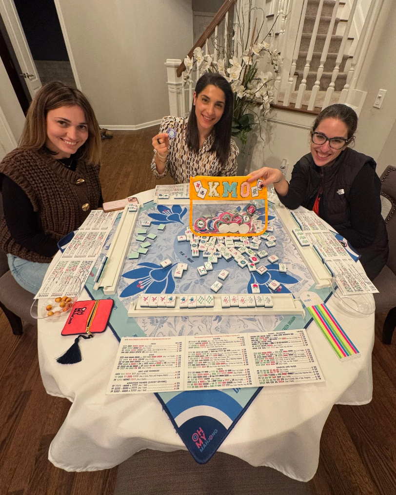 Three women playing Mahjong at a round table with Mahjong tiles, score sheets, and colorful markers, smiling at the camera.