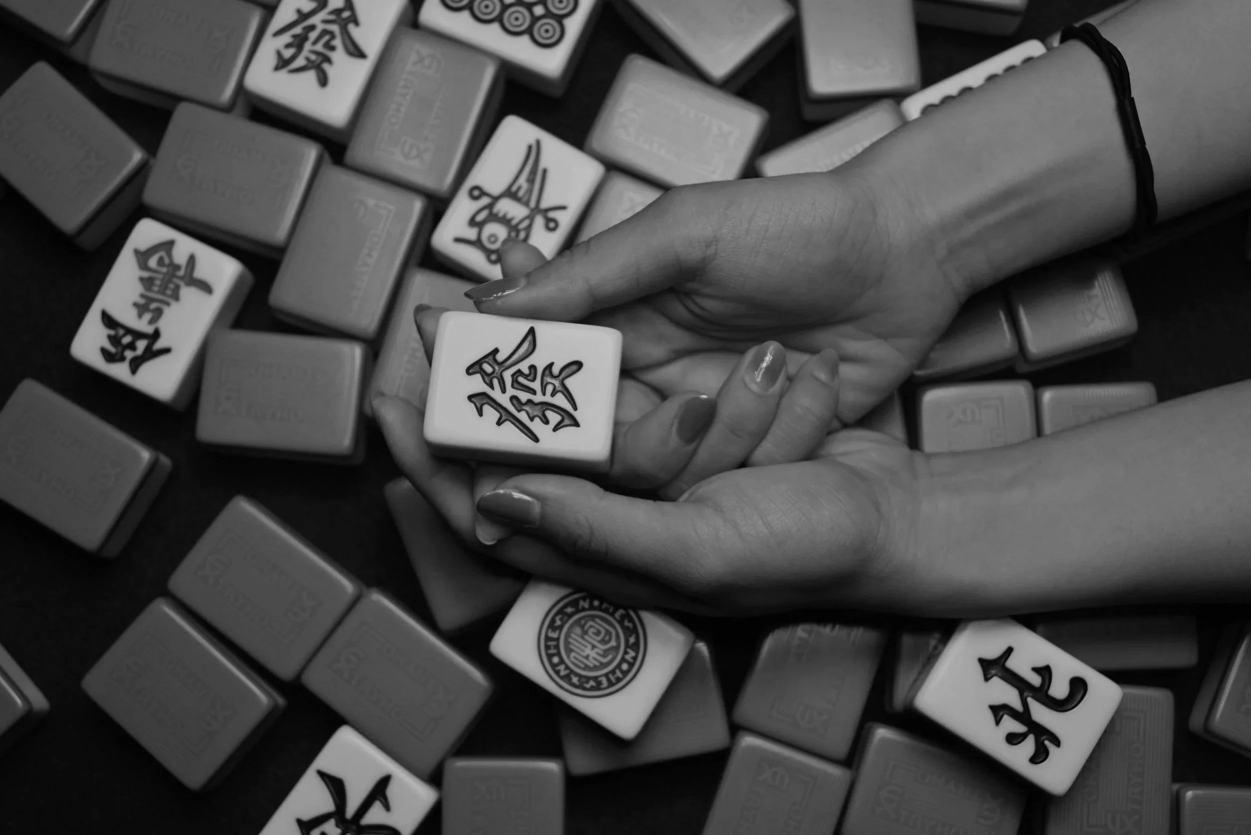 A hand holding a Mah Jong tile with Chinese characters, surrounded by other Mah Jong tiles on a dark surface.