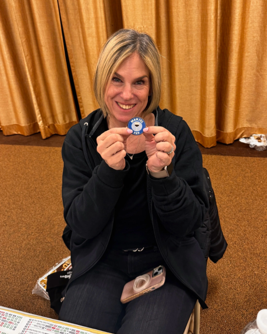 A smiling woman with blonde hair sitting on the floor, holding a blue and white badge that reads "MAJORS TOY." She is wearing a black jacket and has a smartphone with a pop socket on her lap. Behind her is a golden curtain, and on the floor are some items including a plastic bag with chips, a piece of paper, and a white container.