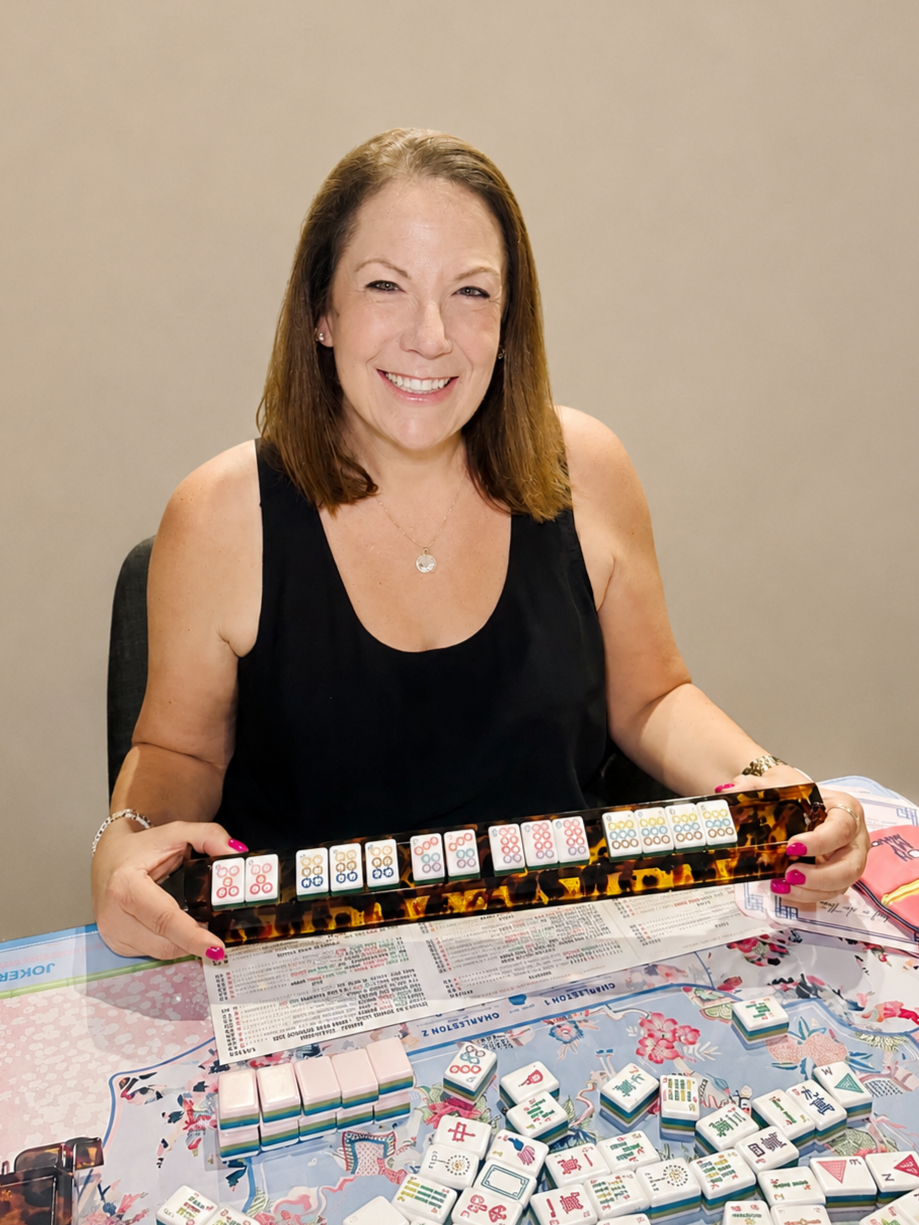 A woman playing Mahjong, sitting at a table with Mah Jong tiles and score sheets, holding a tray of Mah Jong tiles, smiling at the camera.