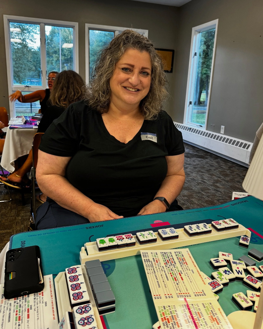 A woman smiling at a mahjong game table, wearing a black shirt with a name tag that reads 'Gail', in a well-lit room with large windows and other people in the background.