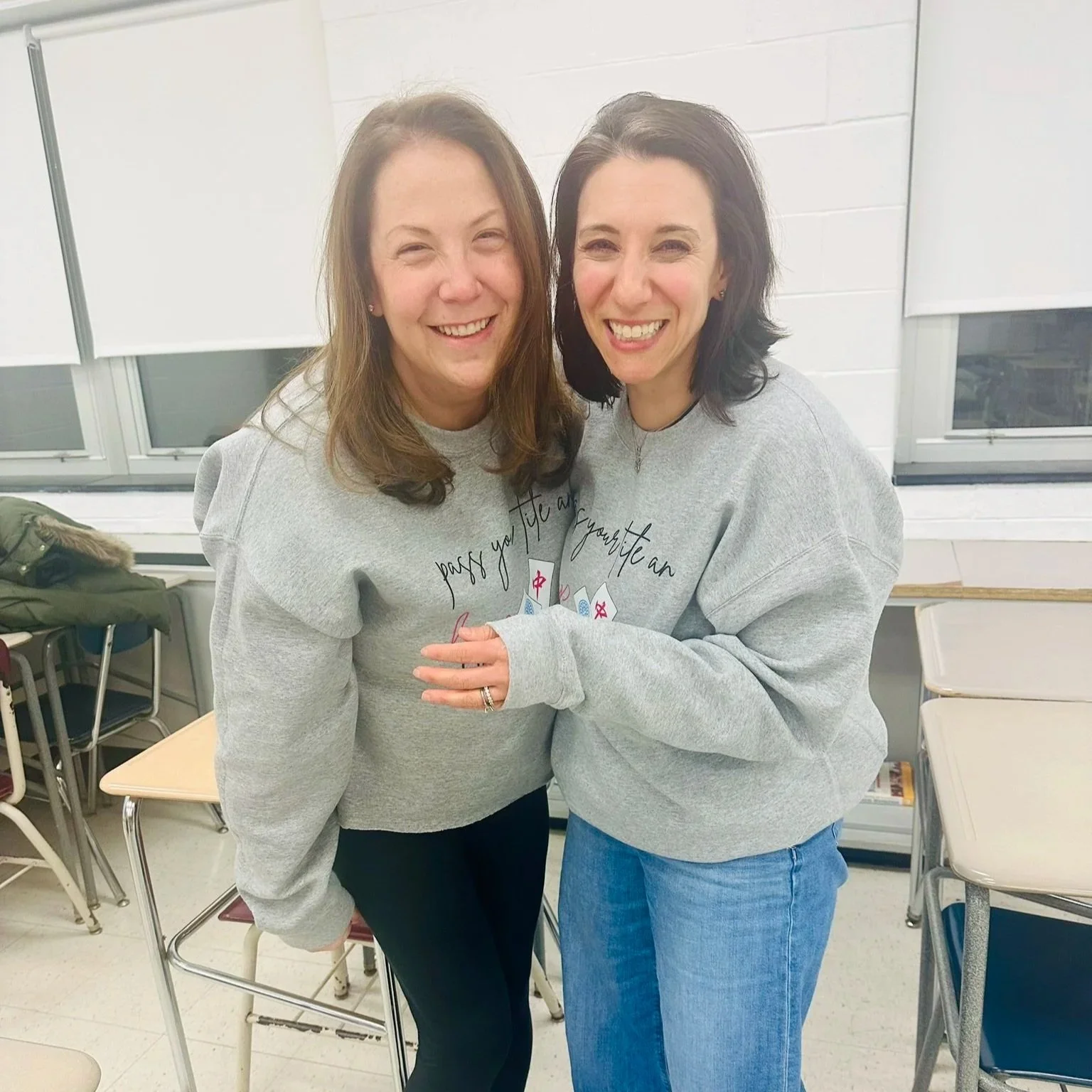 Two women are standing close together in a classroom, smiling and hugging. They are wearing gray sweatshirts with writing and small design on the front. The classroom has white brick walls, windows, desks, and chairs in the background.