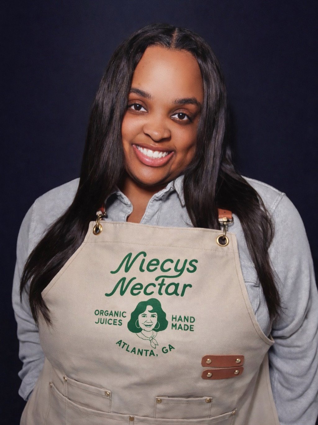 A woman with long dark hair smiling, wearing a grey shirt and a beige apron with green text and illustration, standing against a dark background.
