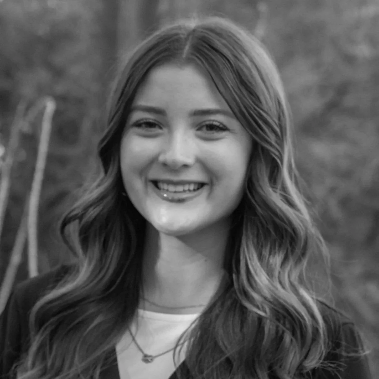 A smiling young woman with long wavy hair, wearing a dark jacket and a necklace, outdoors with blurred nature background in black and white.
